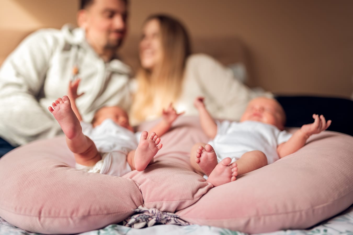 Bliźniaczki leżą na łóżku, w tle rodzice się przytulają — sesja noworodkowa bliźniąt Gdynia