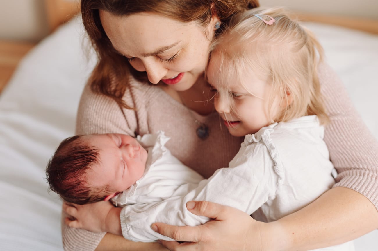 Mama z córkami i noworodkiem przytulają się na łóżku — domowa sesja noworodkowa Gdynia