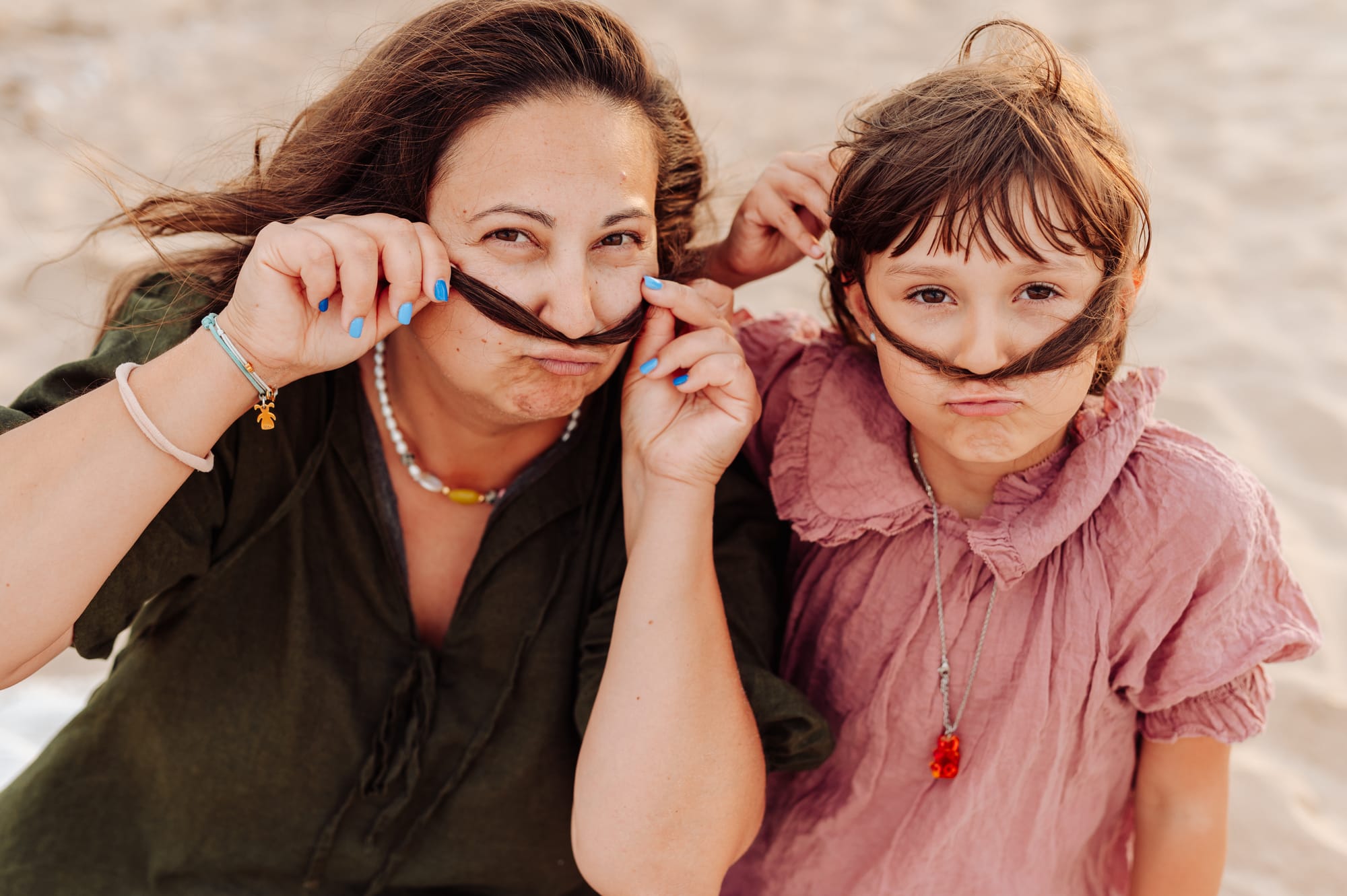 Mama i córka podczas zachodu słońca na plaży — robią sobie wąsy z włosów i śmieją się