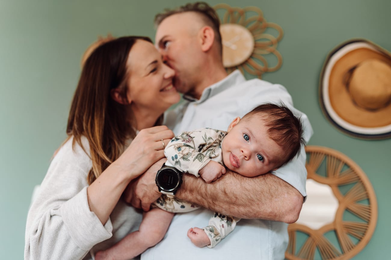 Tata całuje mamę mając niemowlaka na ręce naturalna niemowlęca sesja lifestyle