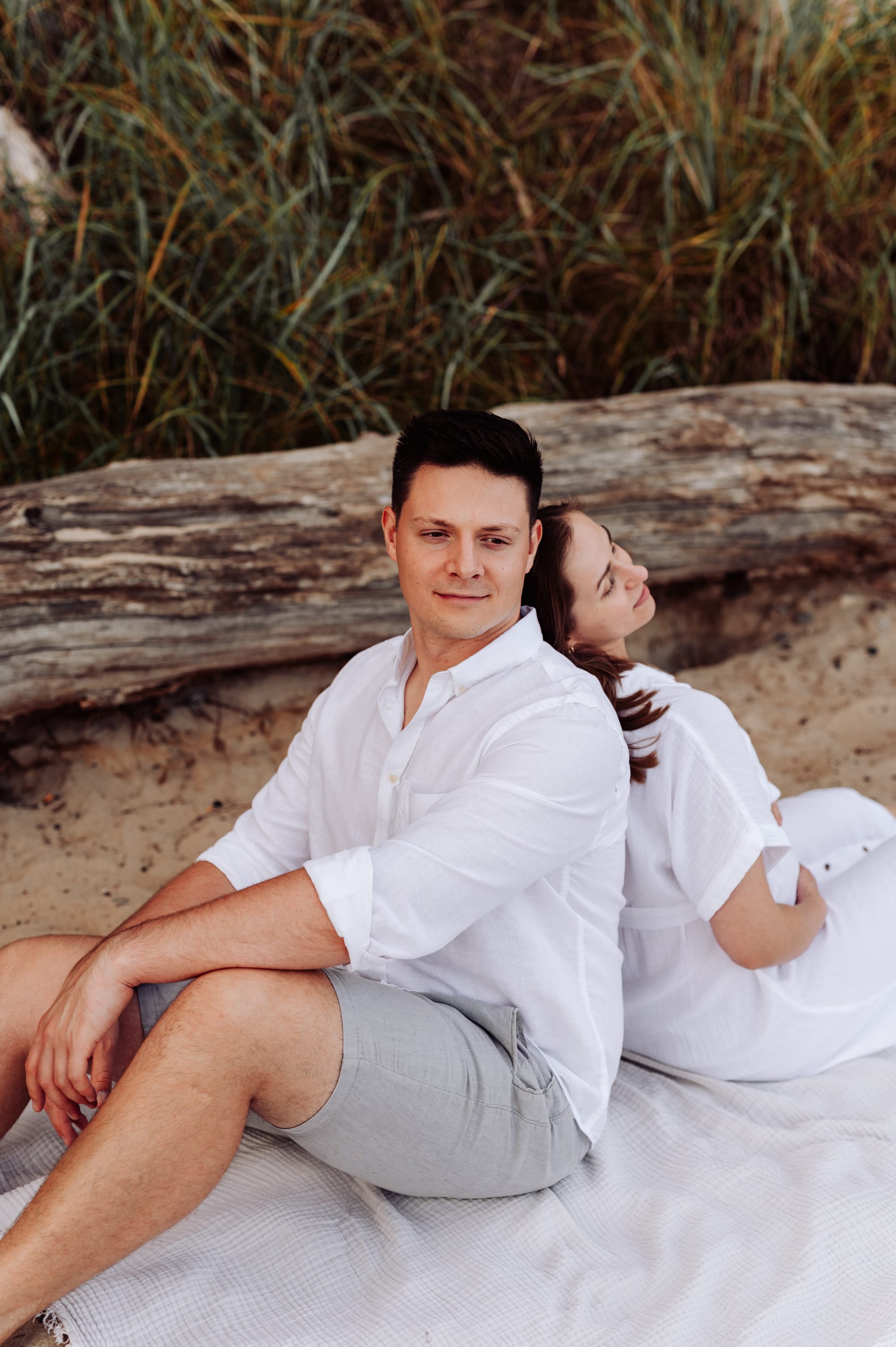 Przyszły tata siedzi na plaży plecami do partnerki i wpatruje się w morze — sesja ciążowa Gdynia, fotograf Natalia Kobylak