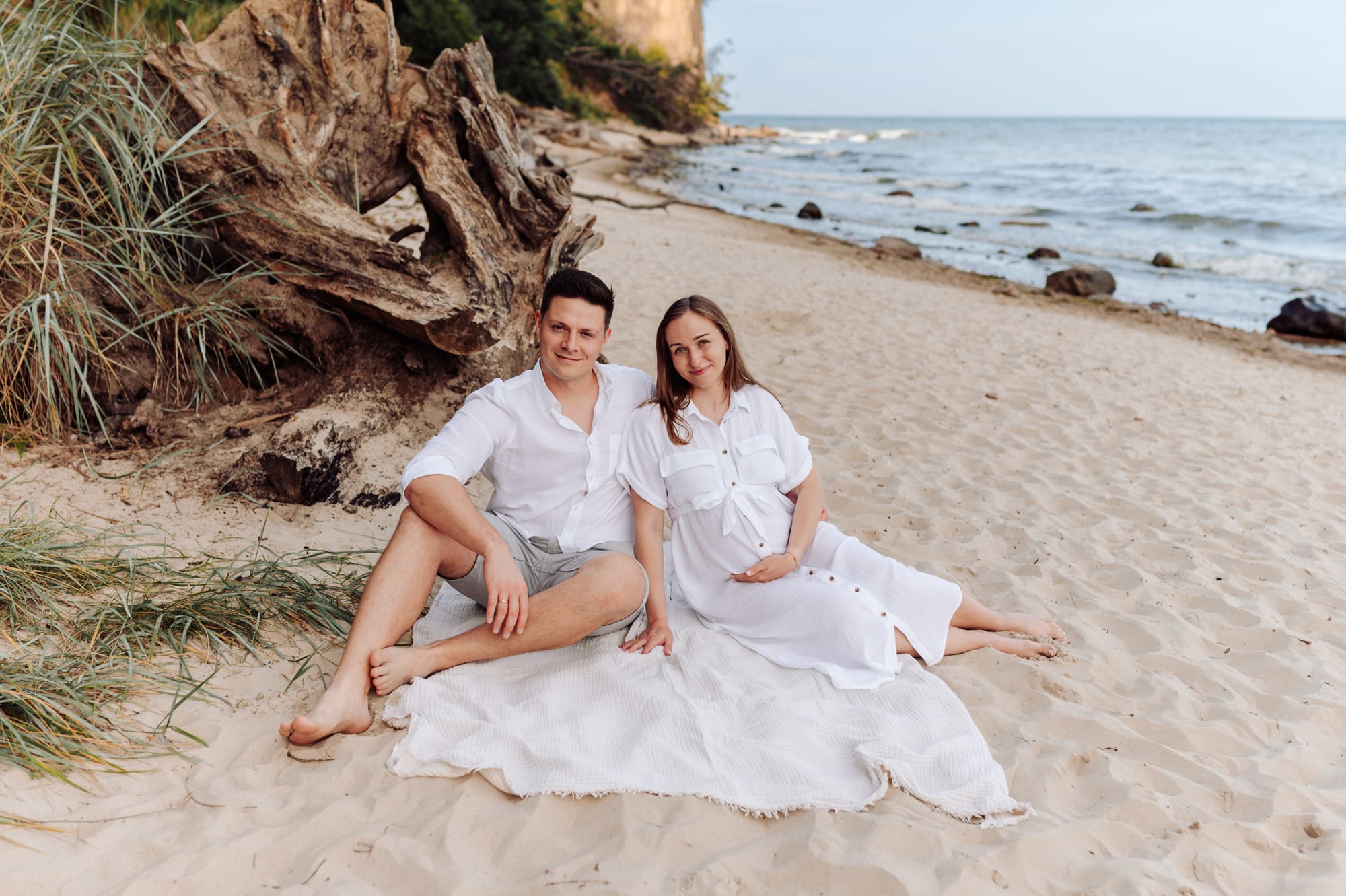 Para siedzi na kocu na plaży w Orłowie, w tle klif, uśmiechnięci — sesja ciążowa Gdynia, fotograf Natalia Kobylak