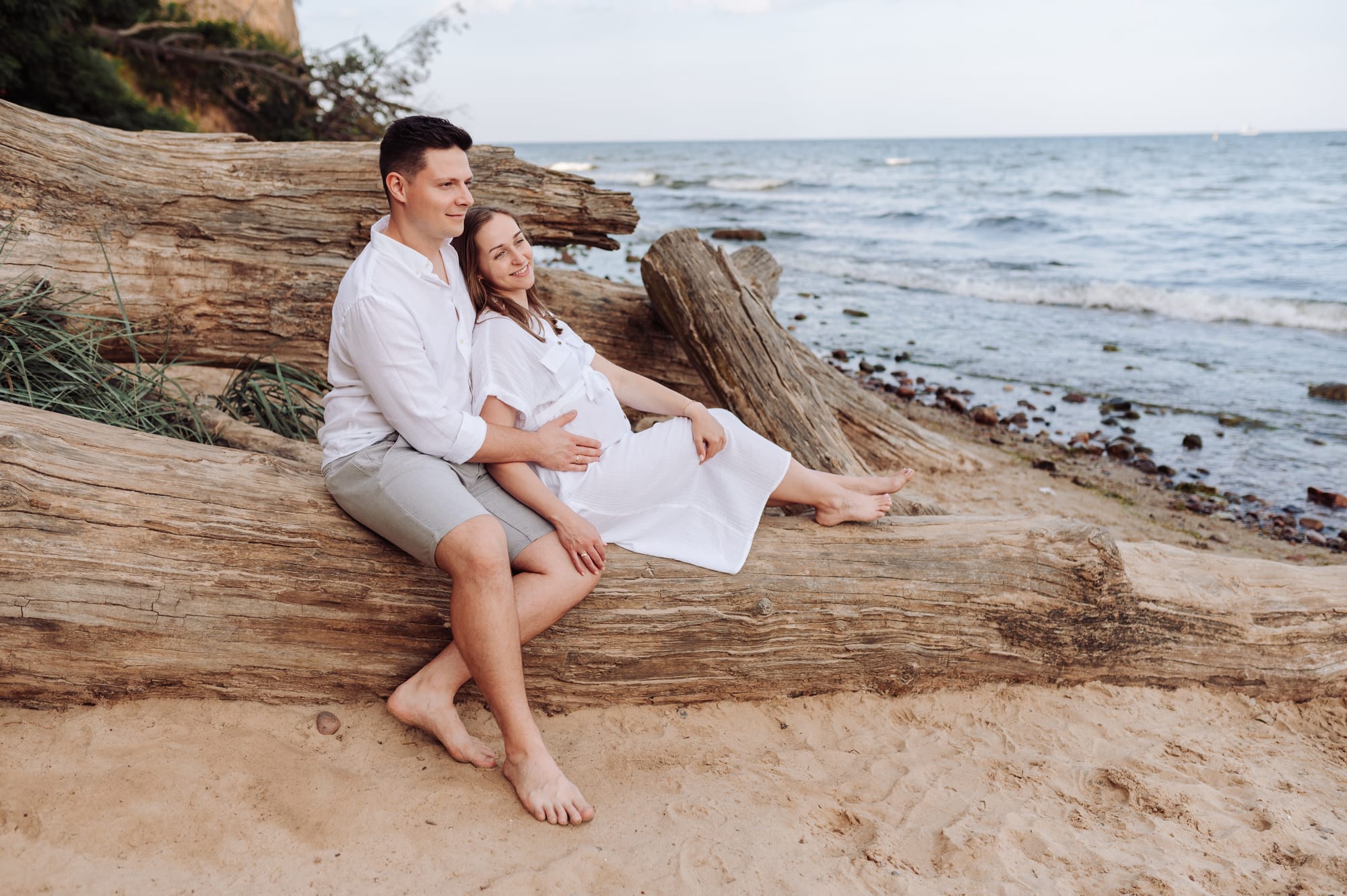 Przyszli rodzice siedzą razem na konarze drzewa i wpatrują się w morze — sesja ciążowa Orłowo Gdynia, fotograf Natalia Kobylak