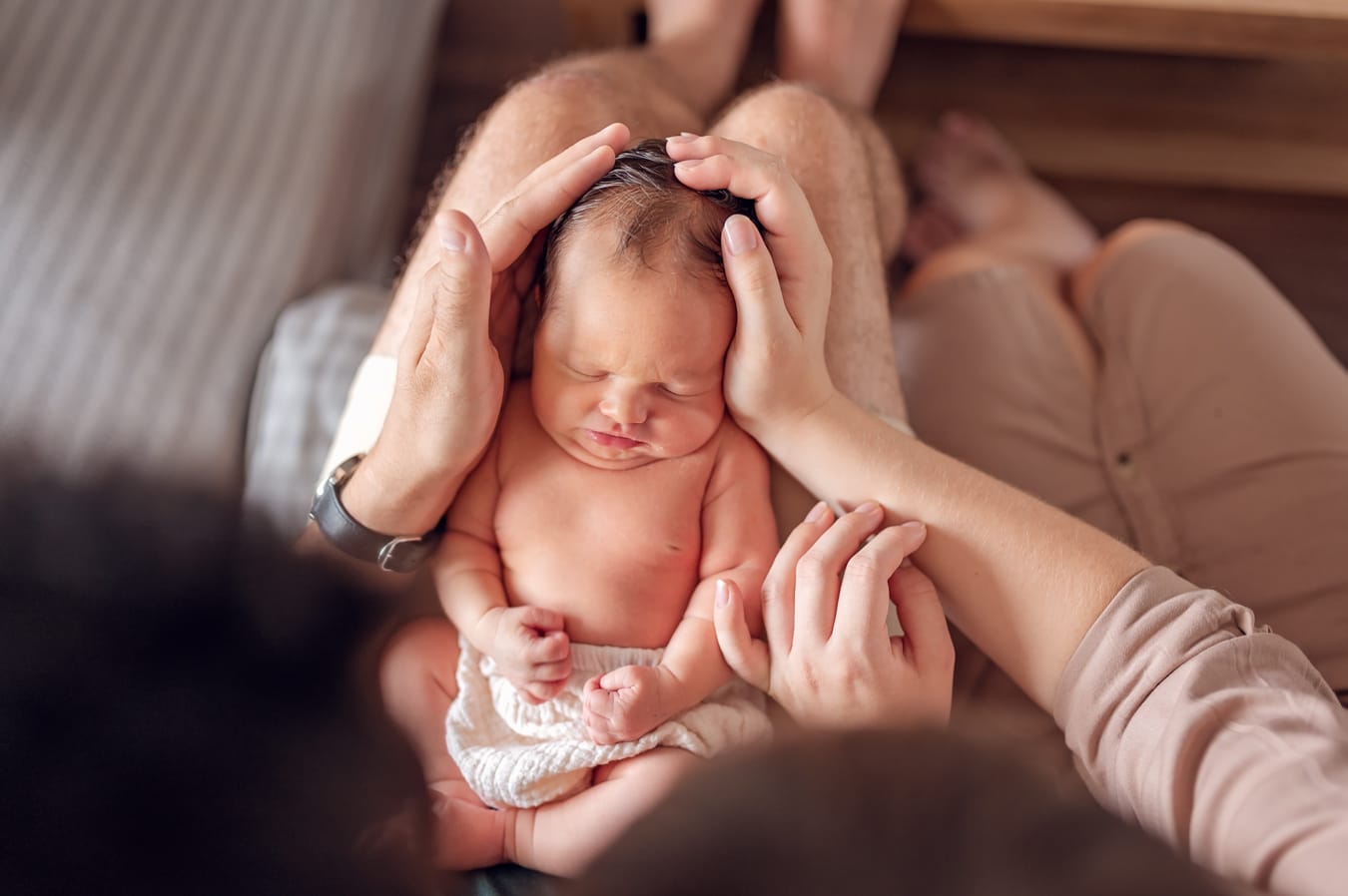 Noworodek śpiący na kolanach taty otulony dłońmi rodziców. 