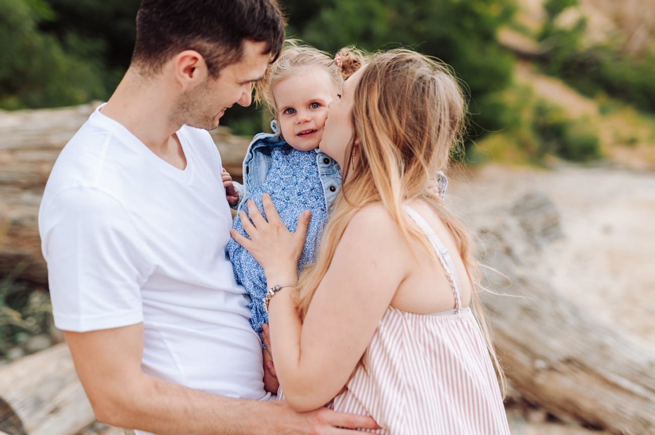 Mama całuje córkę na rękach taty sesja ciążowa plaża