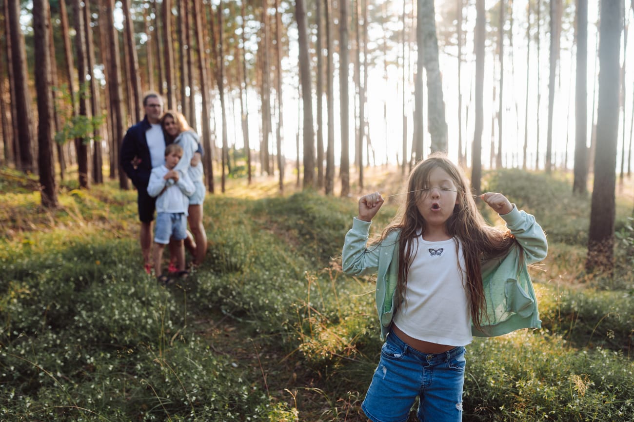 Dziewczynka bawiąca się w lesie a jej rodzina w tle przytula się sesja rodzinna