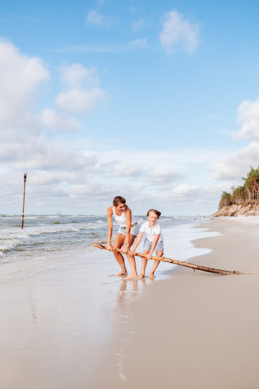 Synek z mamą ciagną pień drzewa po plaży sesja rodzinna