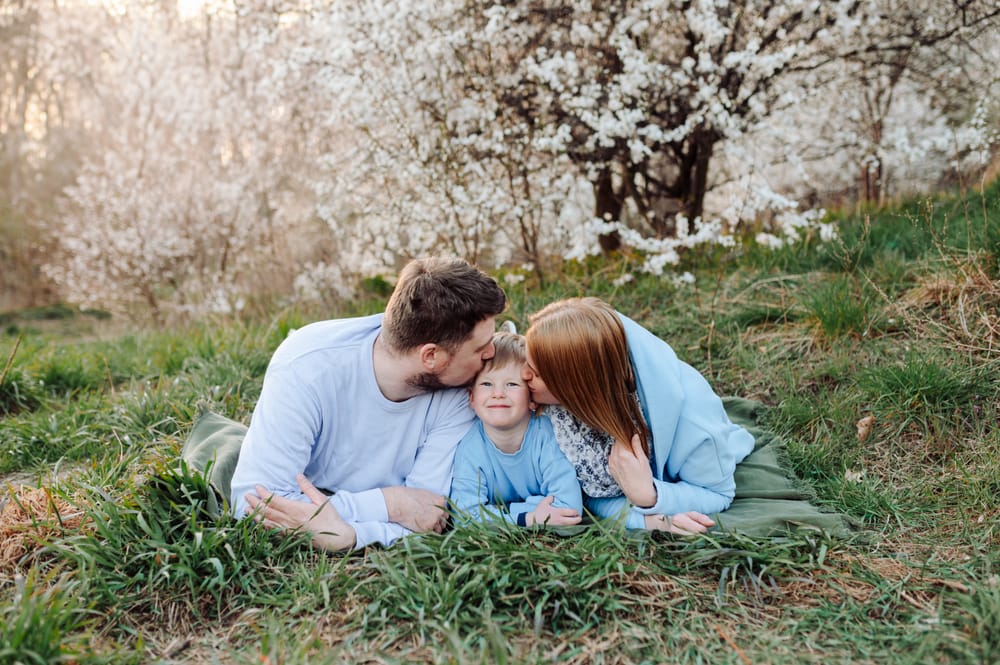 Rodzice całują uśmiechniętego synka na tle kwitnących wiosennych drzew z promieniami zachodzącego słońca — fotografia dziecięca Gdynia
