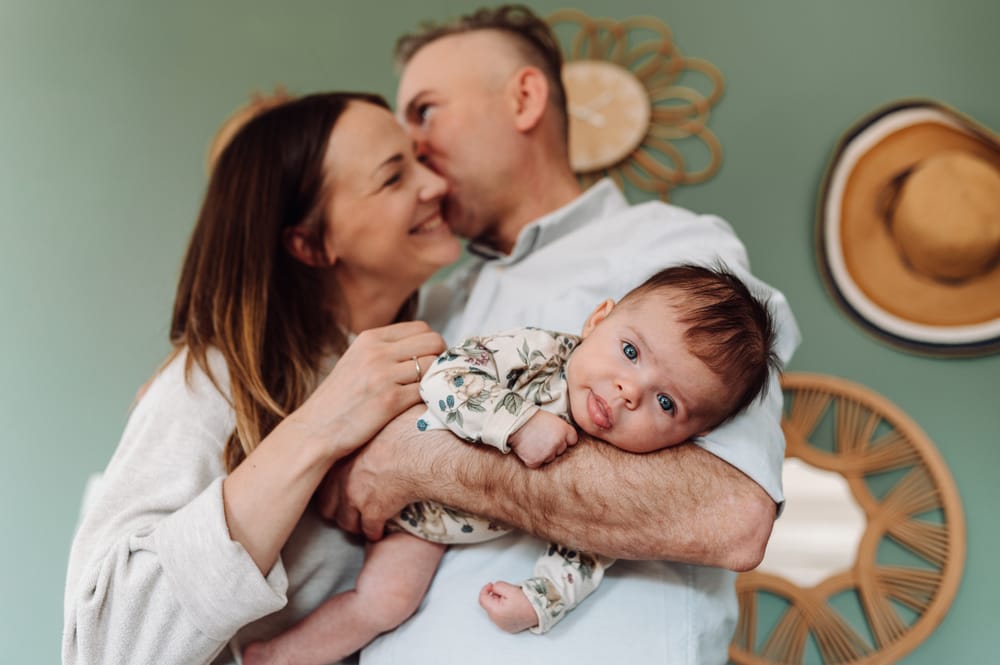 Córeczka na rękach taty wpatrzona w obiektyw, w tle rodzice się przytulają — sesja noworodkowa Gdynia Trójmiasto