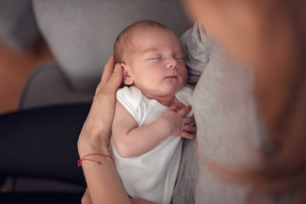 Noworodek spokojnie śpiący w ramionach mamy w trakcie profesjonalnej sesji noworodkowej w domu.