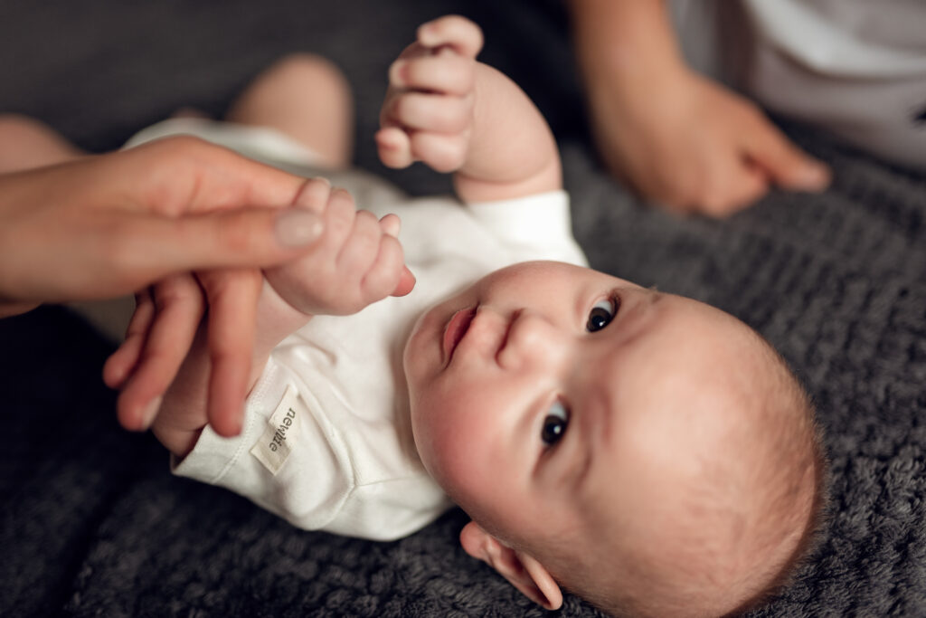 Niemowlę trzyma mamę za palec leżąc na łóżku w domowym zaciszu.