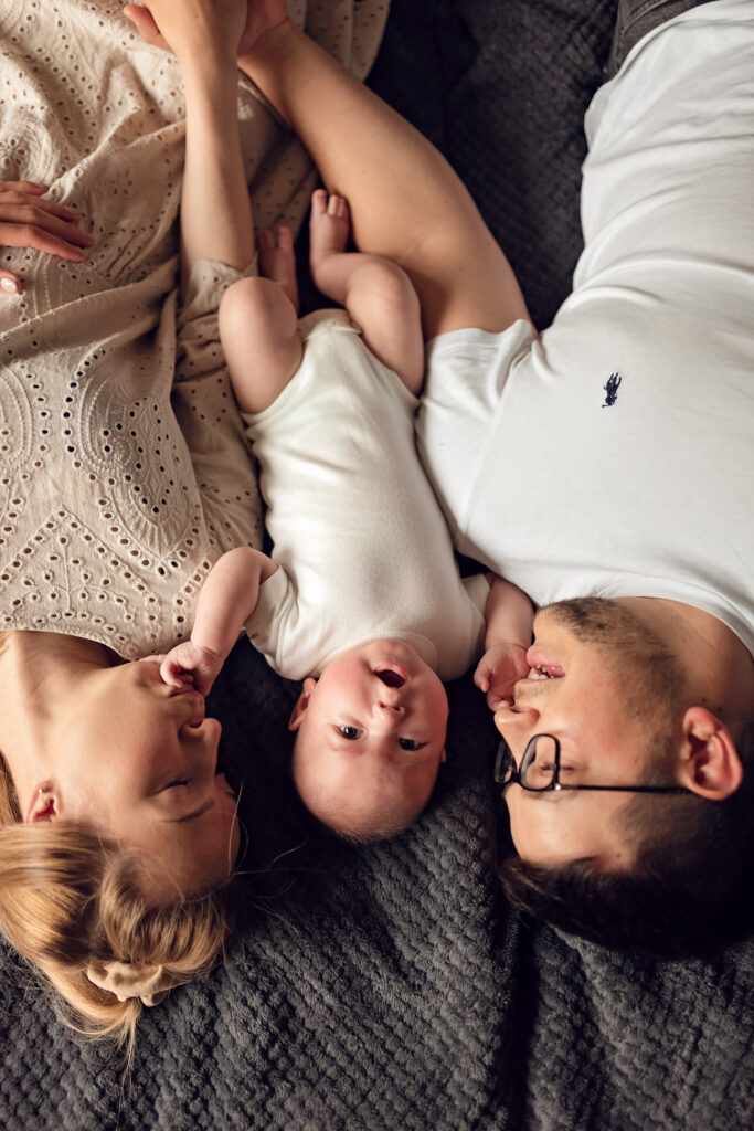 Niemowlę lezące między rodzicami i wyciągające do nich dłonie.