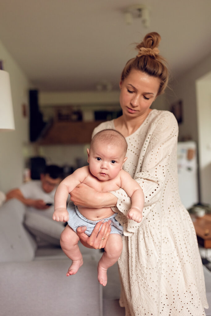 Mama bujająca niemowlę na rękach a w tle tata przeglądający wiadomosci na telefonie. 