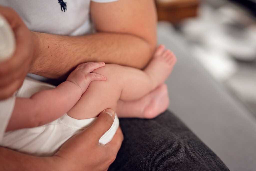 Niemowle trzymające dłoń na kolanie w trakcie karmienia butelką. 