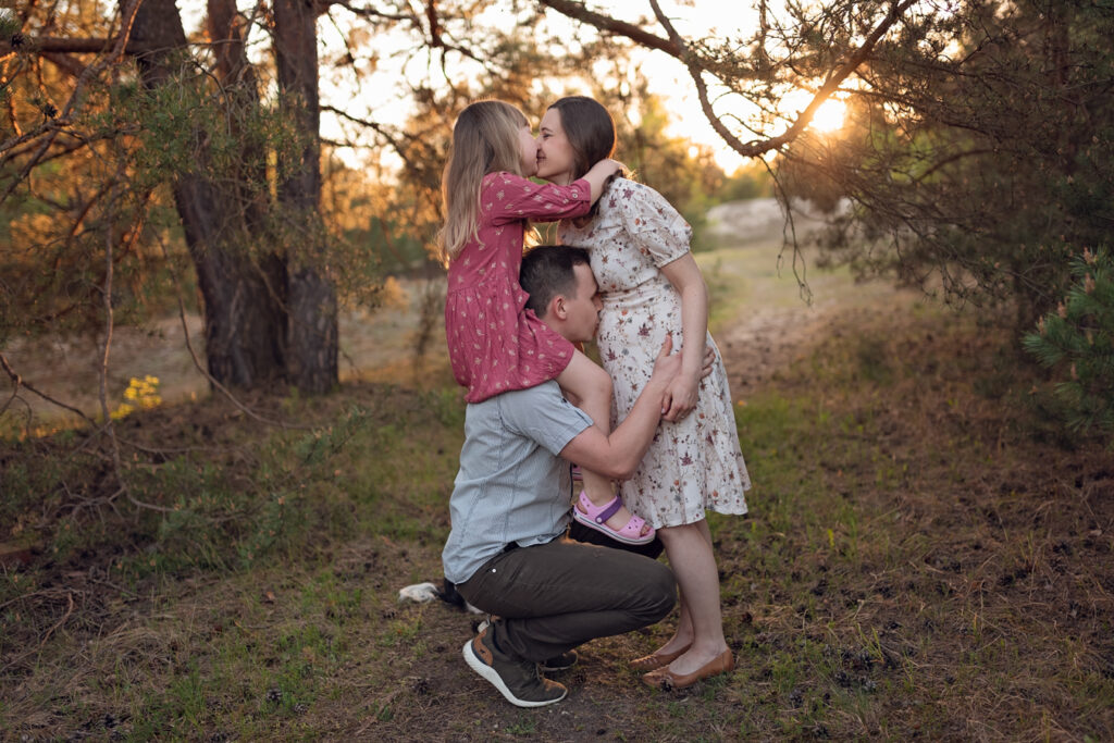 Mama całuje córkę, a tata całuje rosnący brzuszek podczas rodzinnej sesji ciążowe.