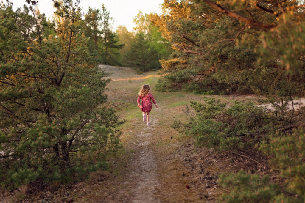 Zabawa wśród drzew – Córka biegnie przed mamą i psem. W tle widać wysokie sosny oświetlone ciepłym światłem zachodzącego słońca.