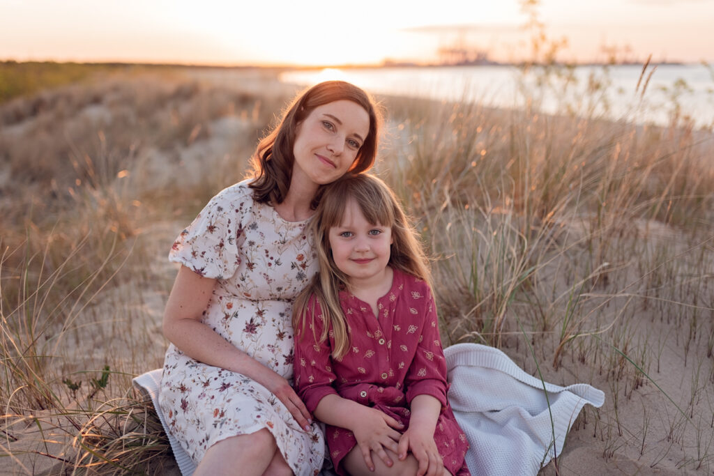Przytulny moment na plaży – Przyszła mama i córka siedzą na piasku,  W oddali widać spokojne morze skąpane w blasku zachodzącego słońca.
