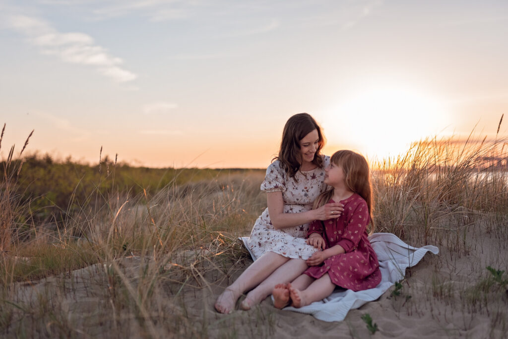 Mama i córka wpatrzone w siebie podczas rodzinnej sesji na plaży z zachodem słońca w tle.