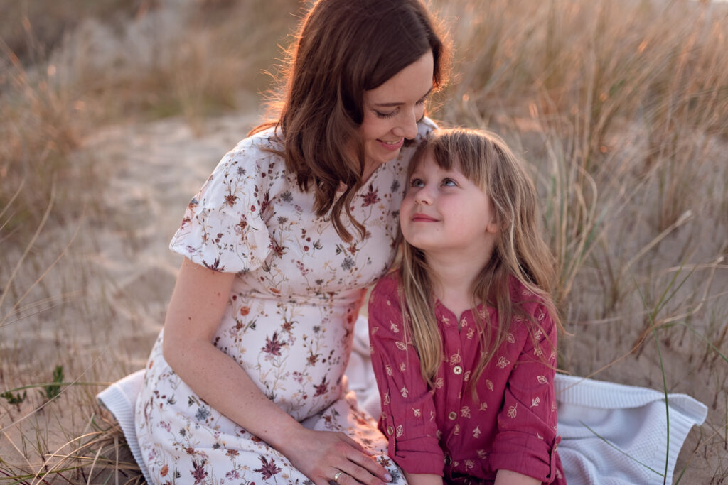Mama w ciąży wpatrzona w starszą córkę otulone promieniami zachodzącego słońca.