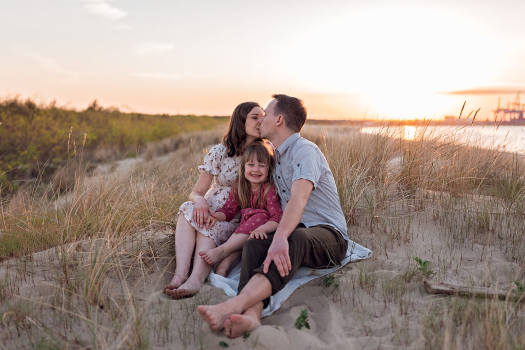 Rodzinna radość w plenerze – Cała trójka – mama, córka i tata – siedzą na plaży a w tle zachodzi słońce.
