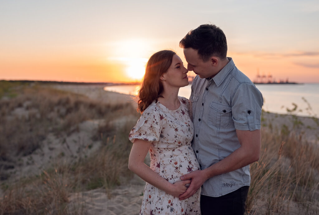 Kobieta w ciąży czule wpatrzona w partnera otuleni promieniami zachodzącego słońca.