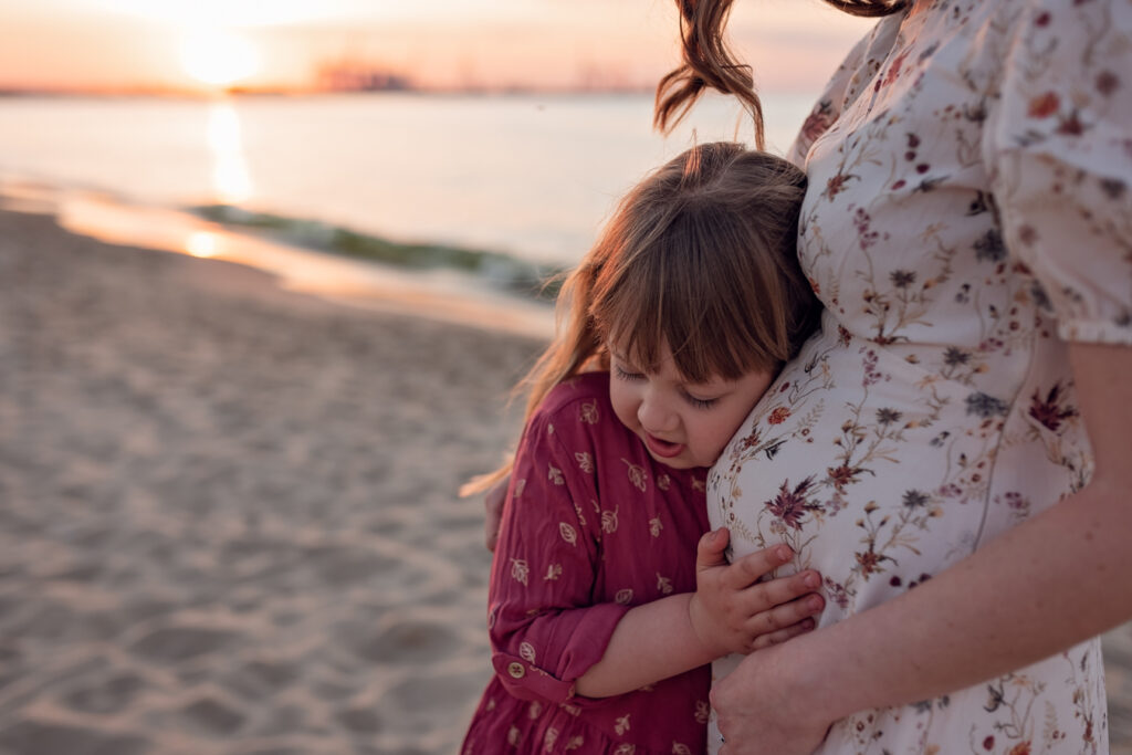 Oczekiwanie na nowego członka rodziny – Mama trzyma dłonie na brzuszku, a córka wsłuchuje się w delikatnie kopnięcia siostry.

