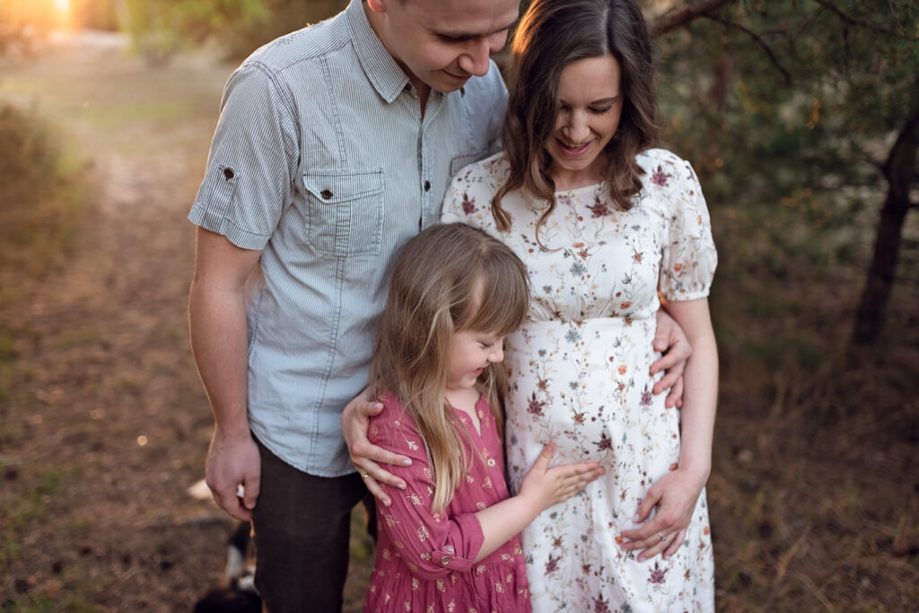 Rodzinny portret na którym dziewczynka przytula ciążowy brzuszek swojej mamy.