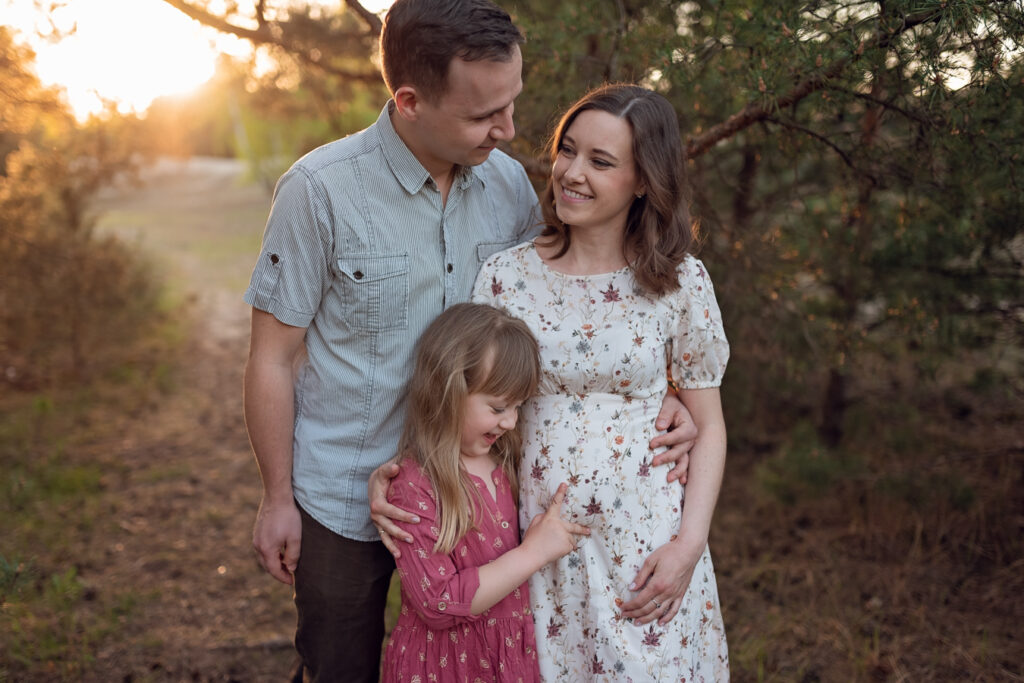Rodzinna chwila w lesie – Mama, córka i pies zatrzymują się wśród wysokich sosen. Córka przytula mamę, a pies radośnie wpatruje się w aparat.