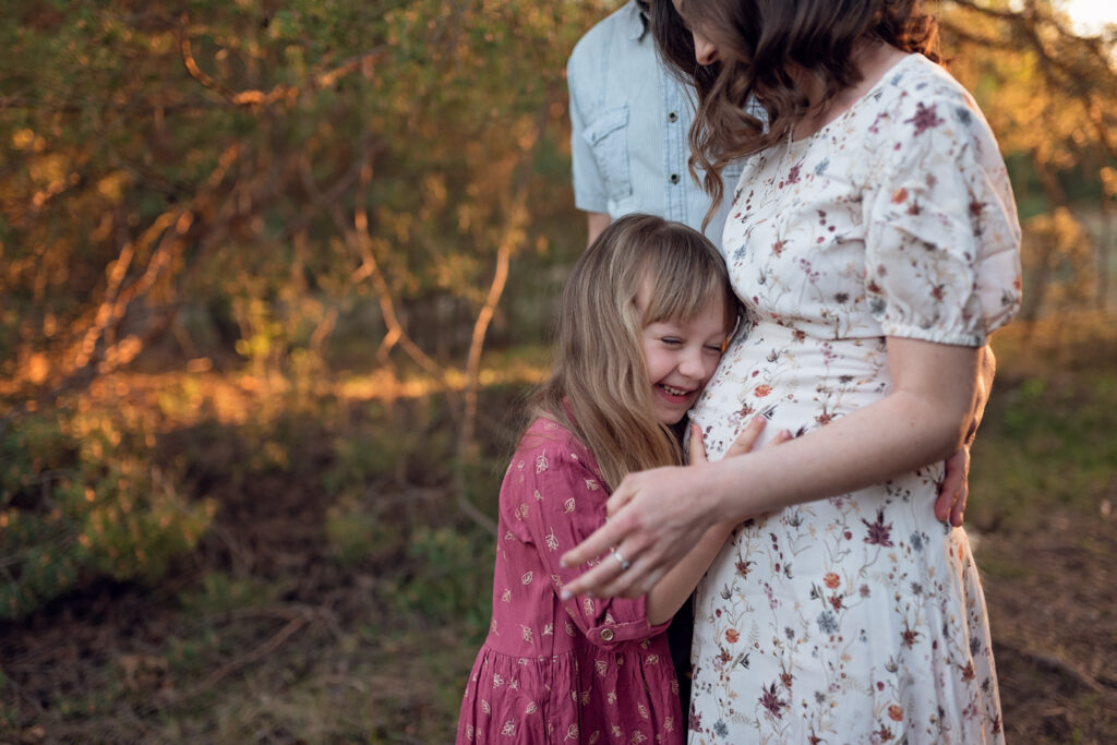 Rodzinne zdjęcie na którym dziewczynka przytula ciążowy brzuszek swojej mamy.