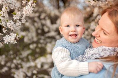 Wiosenna sesja zdjęciowa w Trójmieście - młoda mama trzyma swojego rocznego syna na tle kwitnących drzew.